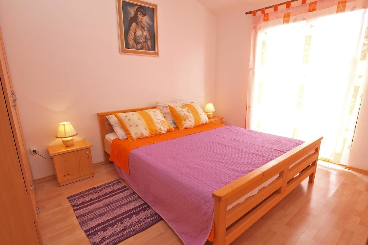 A bedroom with a wooden bed, a purple bedspread, two bedside lamps, a nightstand, a small rug, and a painting on the wall.