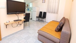 A compact living space with a flat-screen TV mounted on the wall, a sofa with a patterned cushion, a small dining area with a table and chairs, and a kitchenette in the background. - Apartments Lillies 1390 (Fazana)