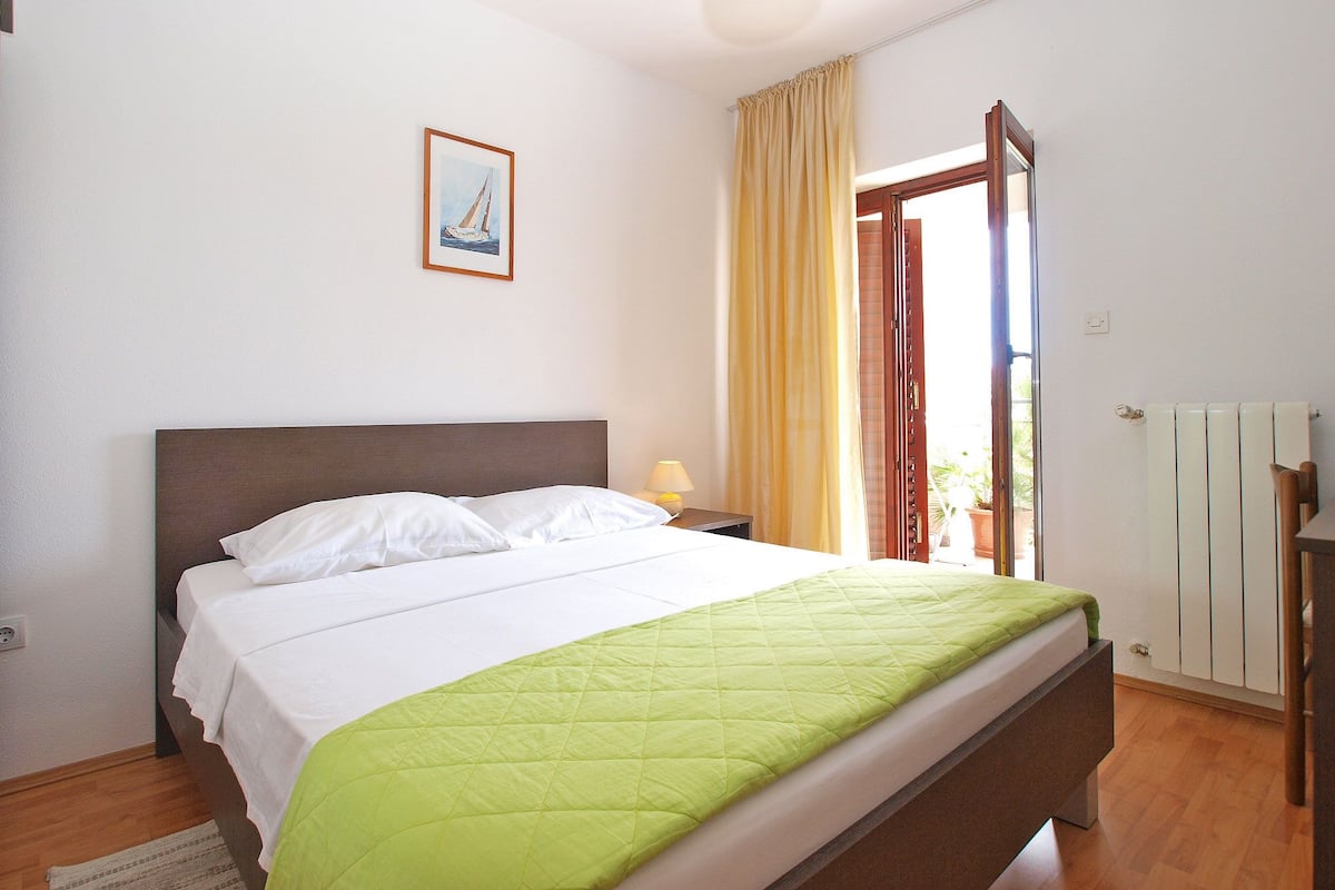 A bedroom with a bed, a nightstand, a lamp, a window with curtains, and a radiator.