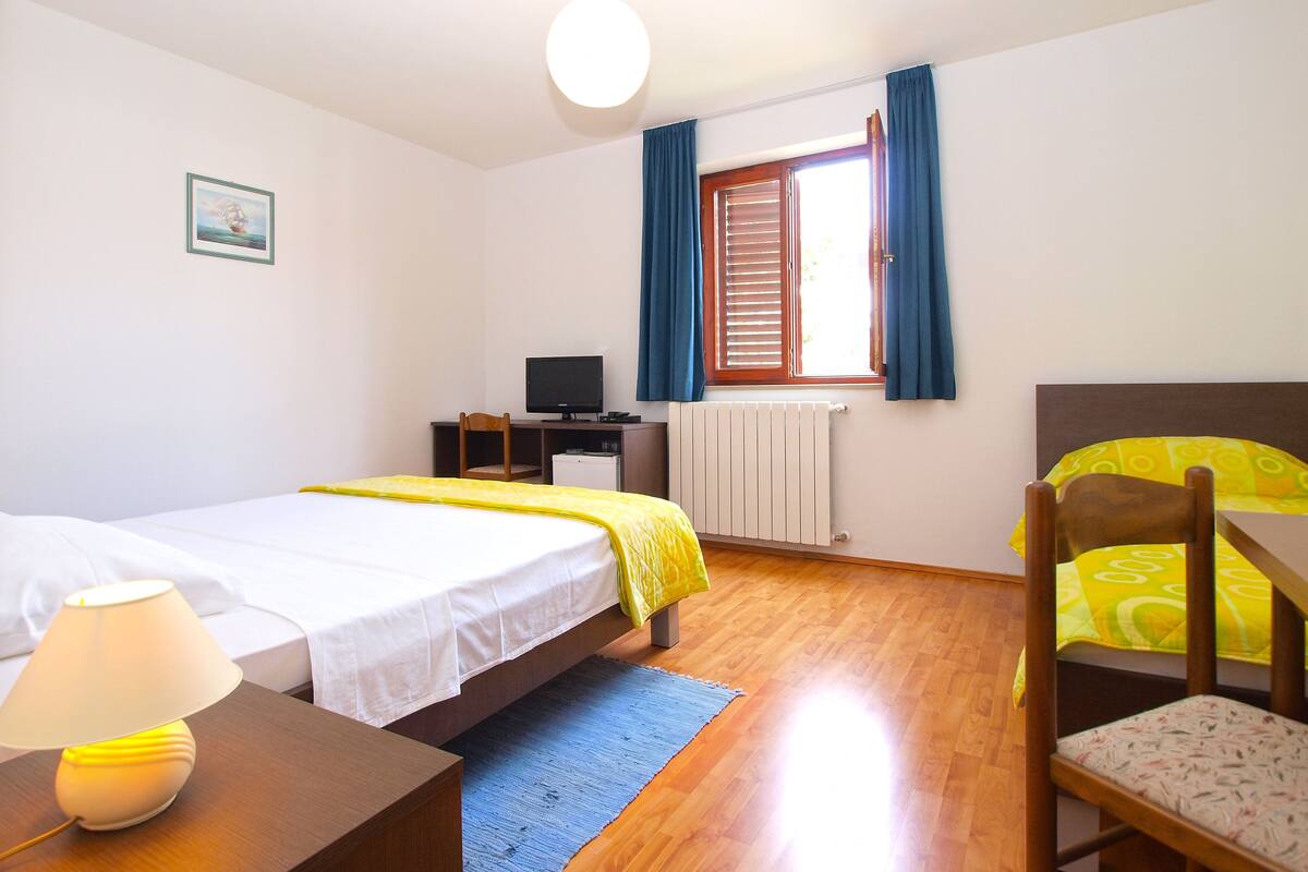 A bedroom with a bed, a desk, a chair, a lamp, a radiator, and a window with curtains.