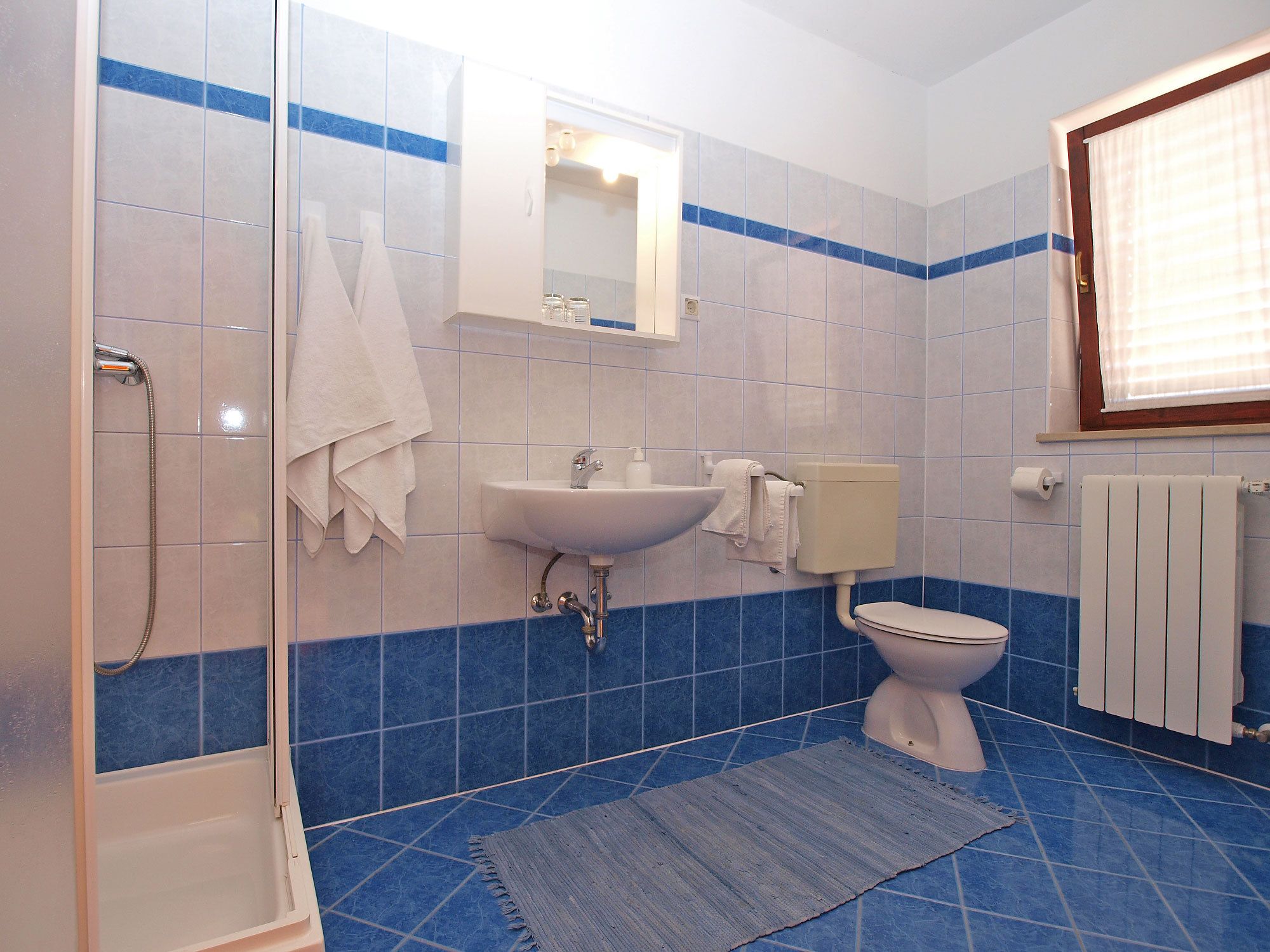 Un baño con ducha, lavamanos, inodoro y ventana con persianas.
