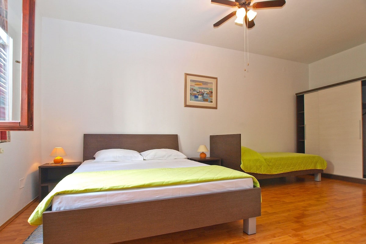 A bedroom with a bed, a nightstand, a lamp, a ceiling fan, a painting, and a wardrobe.