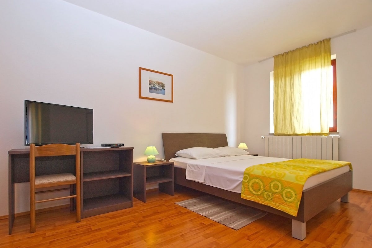 A bedroom with a bed, a desk with a chair, a television, a lamp, and a window with curtains.