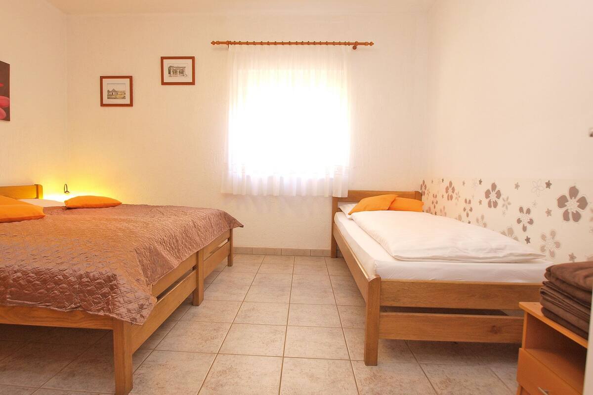 Un dormitorio con dos camas de madera, una ventana con cortinas translúcidas y una lámpara de pared.