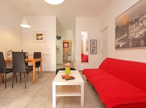 Una sala de estar moderna con un sofá rojo, una mesa de comedor con sillas, una mesa de centro blanca con un jarrón y un cuadro de un paisaje urbano en la pared.