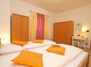 A hotel room with two beds, orange pillows, a wooden wardrobe, a small desk with a mirror, and a lamp. - Apartment Fatima 1514 (Pula)
