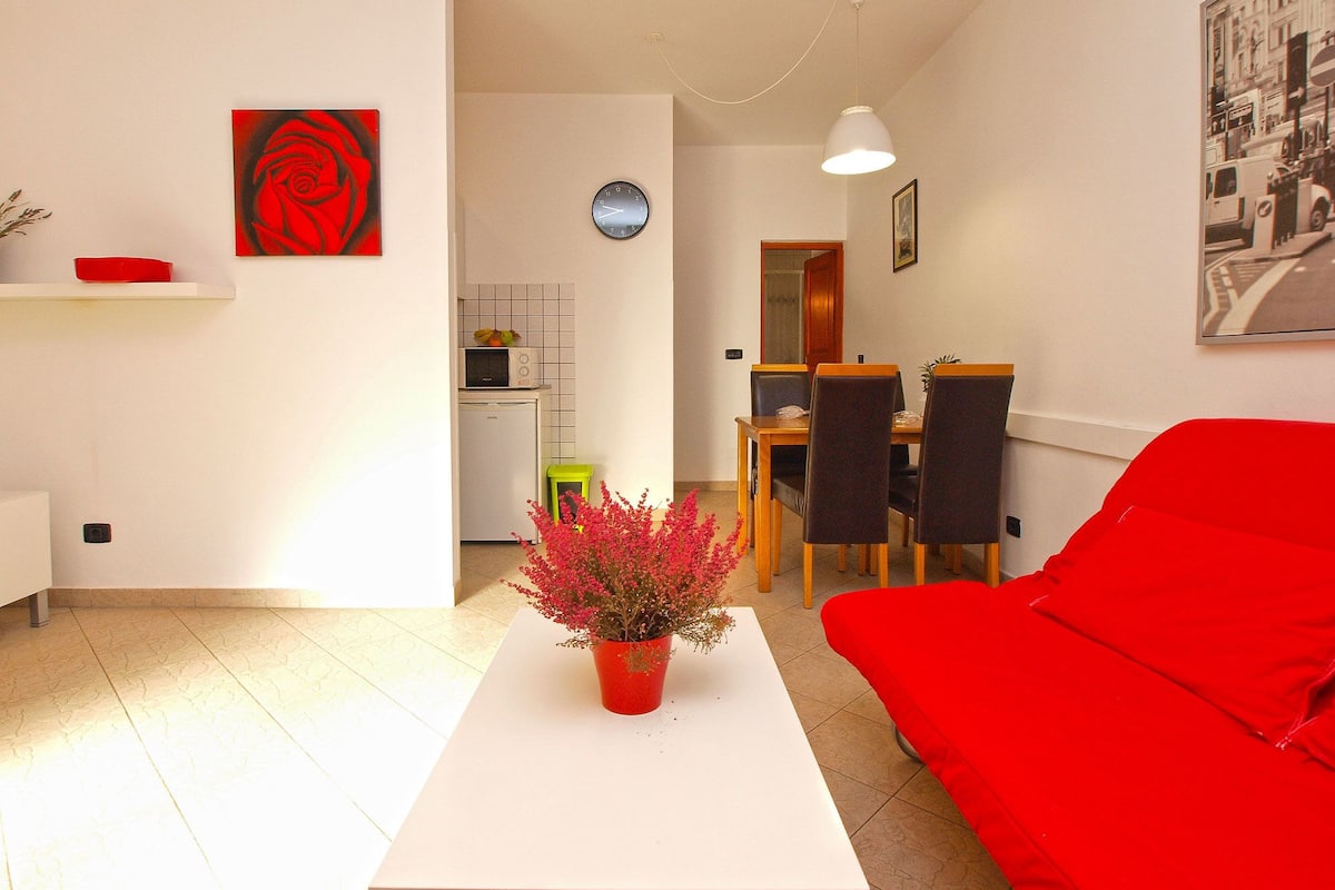 Una sala de estar moderna con un sofá rojo, una mesa de centro blanca con un jarrón de flores rojas, un comedor con una mesa y sillas de madera, y una cocina con microondas y refrigerador.