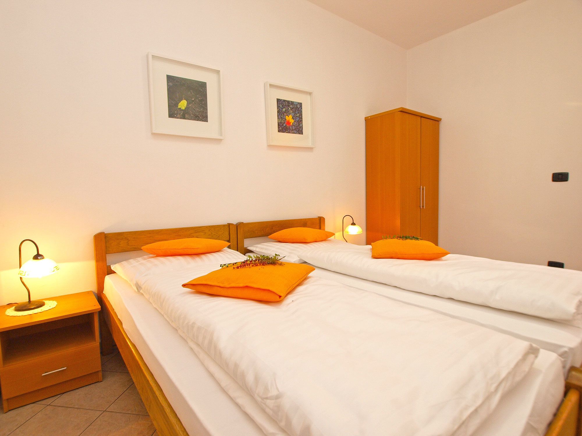 A hotel room with two single beds, a wooden wardrobe, a nightstand with a lamp, and two framed pictures on the wall.