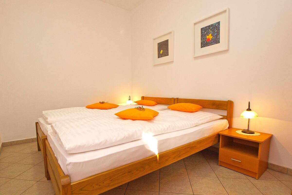 Una cama bien hecha con sábanas blancas y almohadas naranjas, una mesita de noche de madera con una lámpara, y dos cuadros enmarcados en la pared.