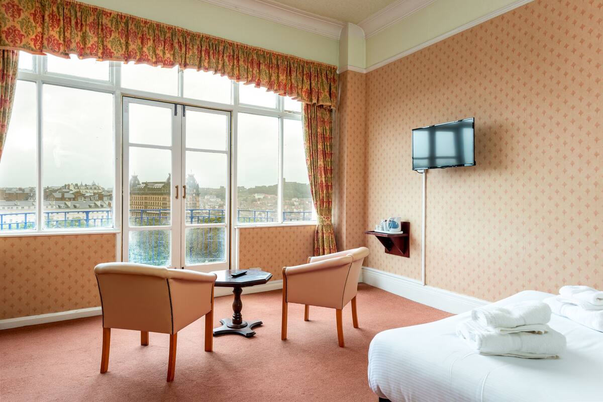 A hotel room with a large window, two chairs, a small table, a bed with neatly folded towels, and a mounted TV.