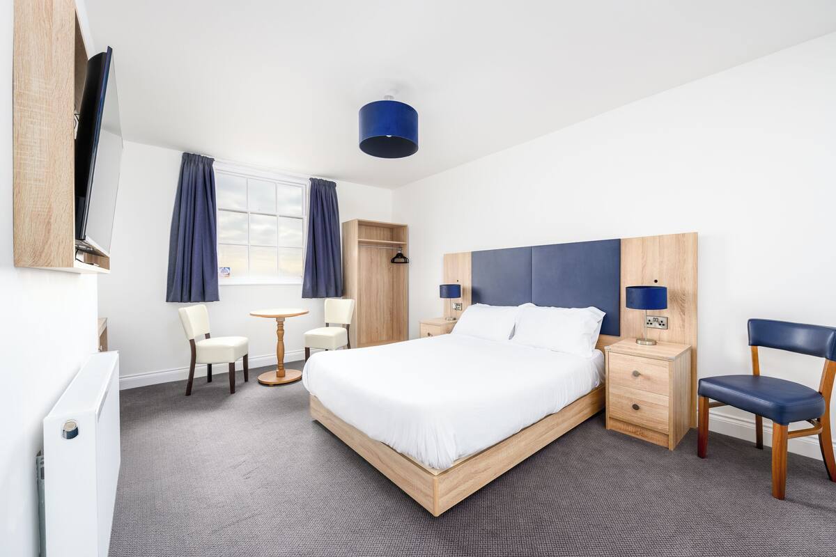 A hotel room with a bed, bedside tables, a desk, chairs, a wardrobe, and a television.