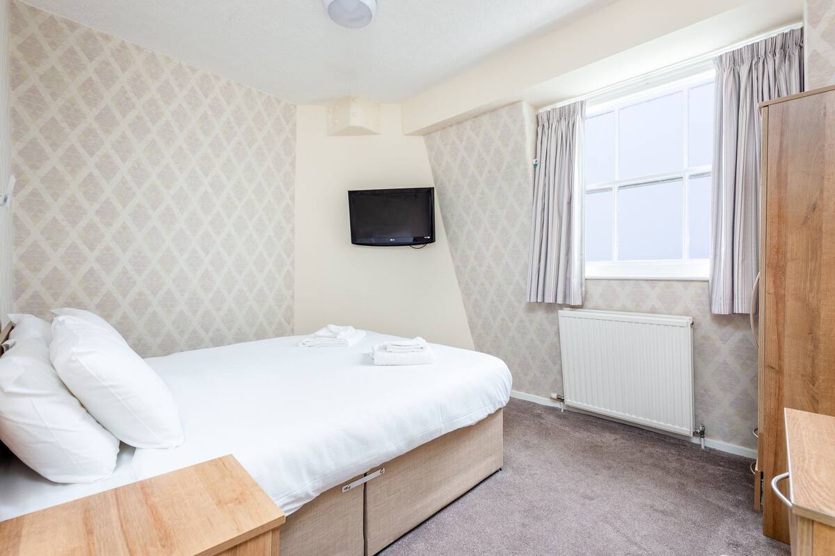 A bedroom with a bed, a television mounted on the wall, a window with curtains, and a radiator.