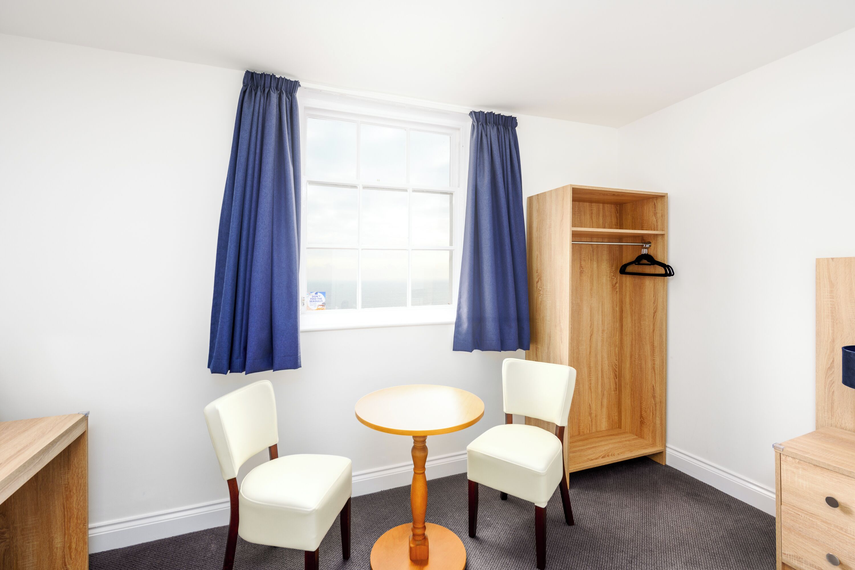 A room with a round wooden table, two white chairs, a wooden wardrobe, and a window with blue curtains.