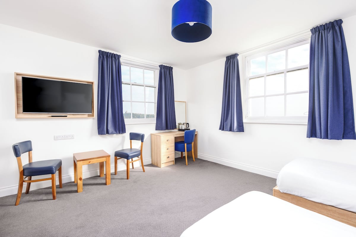 A hotel room with a bed, a desk with two chairs, a television, and large windows with curtains.
