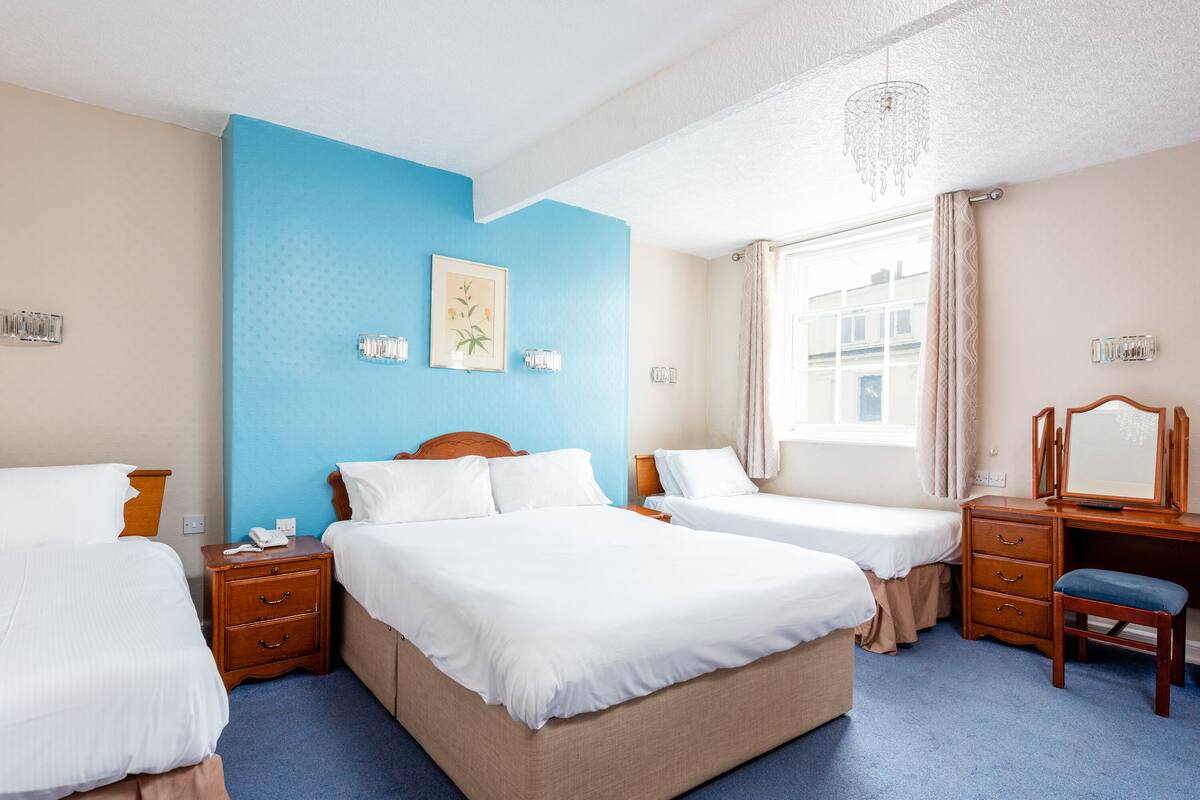 A hotel room with two single beds, a wooden dresser, a desk with a chair, a chandelier, and a window with curtains.