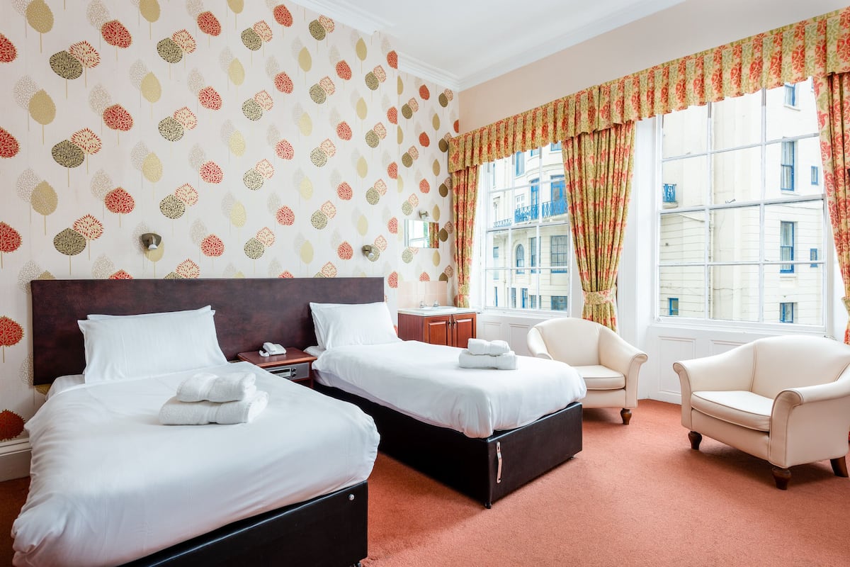 A hotel room with two beds, a sitting area with a chair, a window with curtains, and a patterned wall.