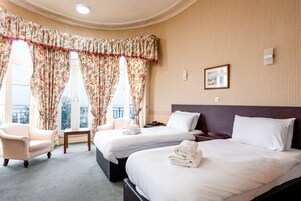 Habitación de hotel con dos camas, una silla, una mesita y un balcón con cortinas.