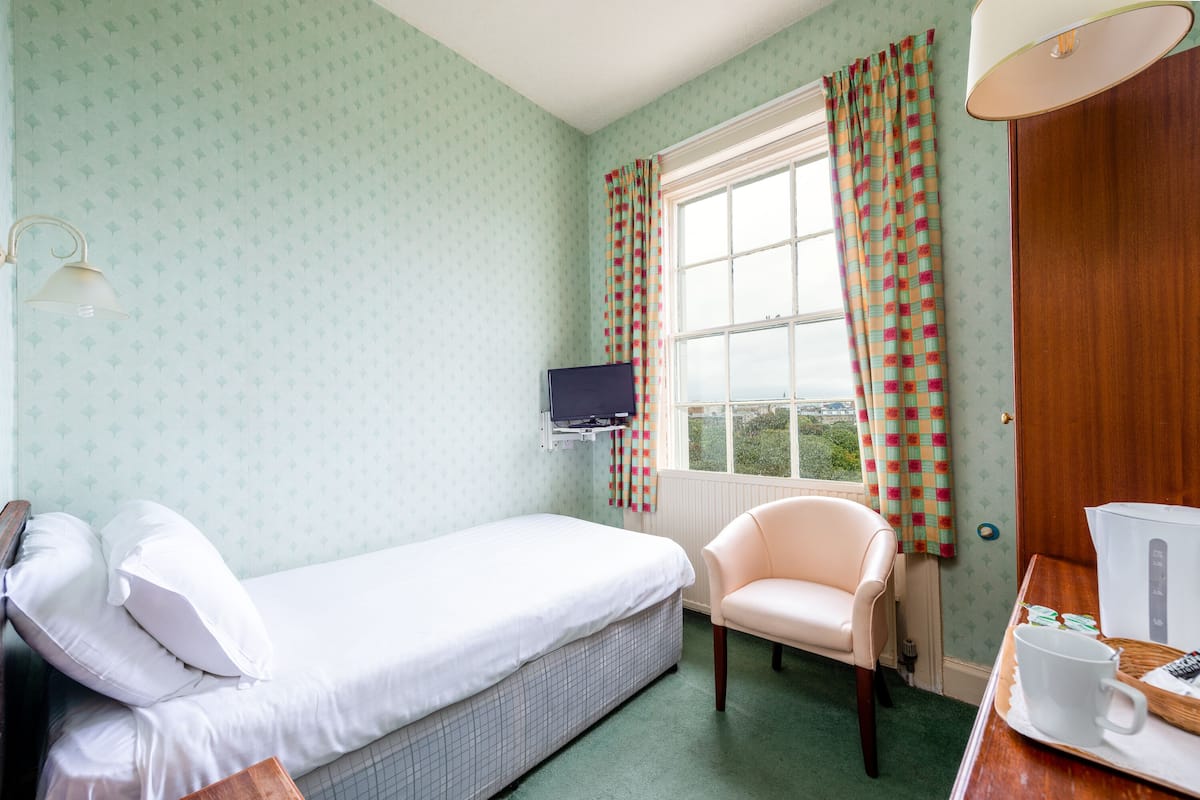 A room with a bed, a chair, a desk with a computer, a window with curtains, and a wall-mounted TV.