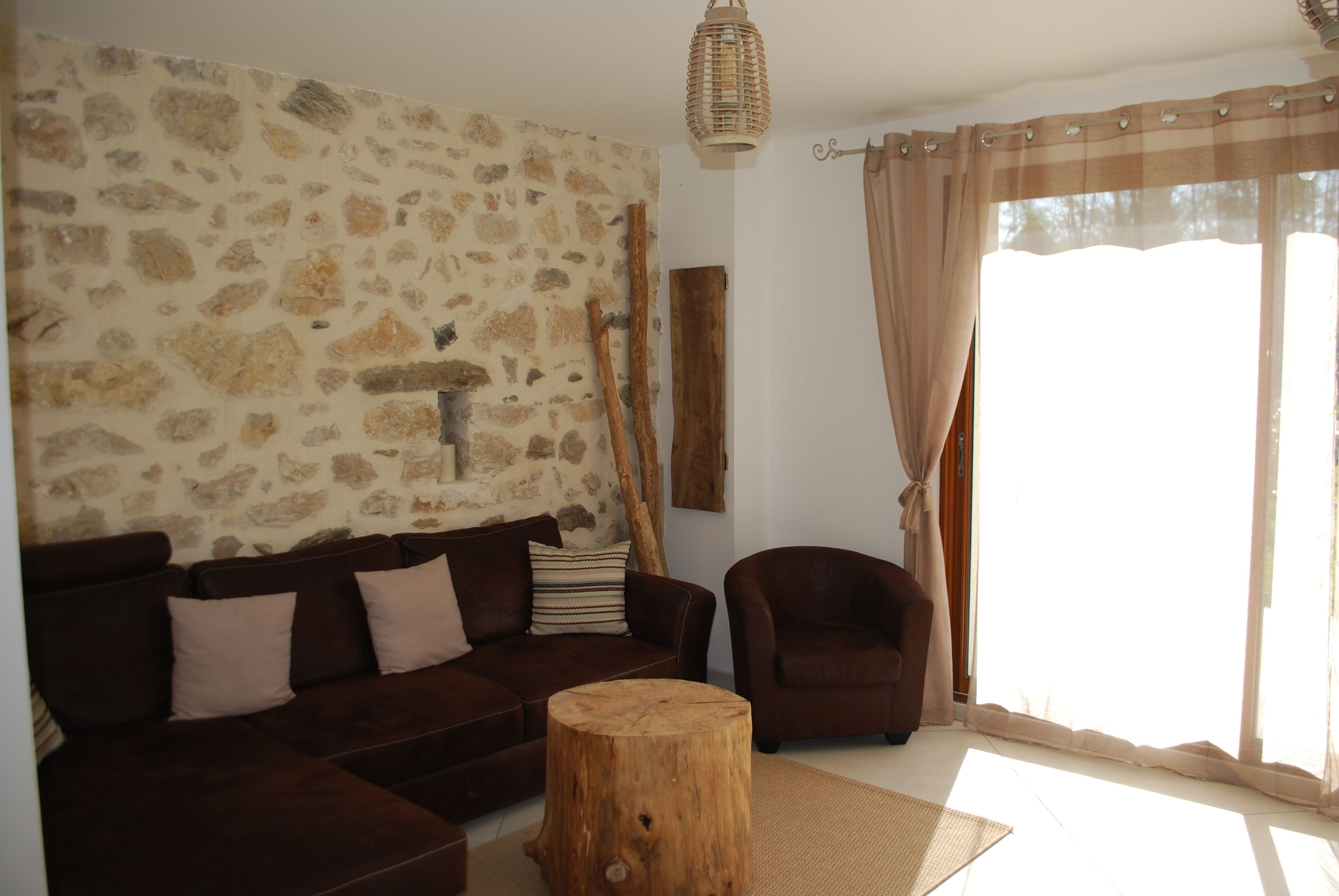 Una sala de estar con una pared de piedra, un sofá marrón, una mesa de centro de madera, un sillón marrón y un ventanal con cortinas.