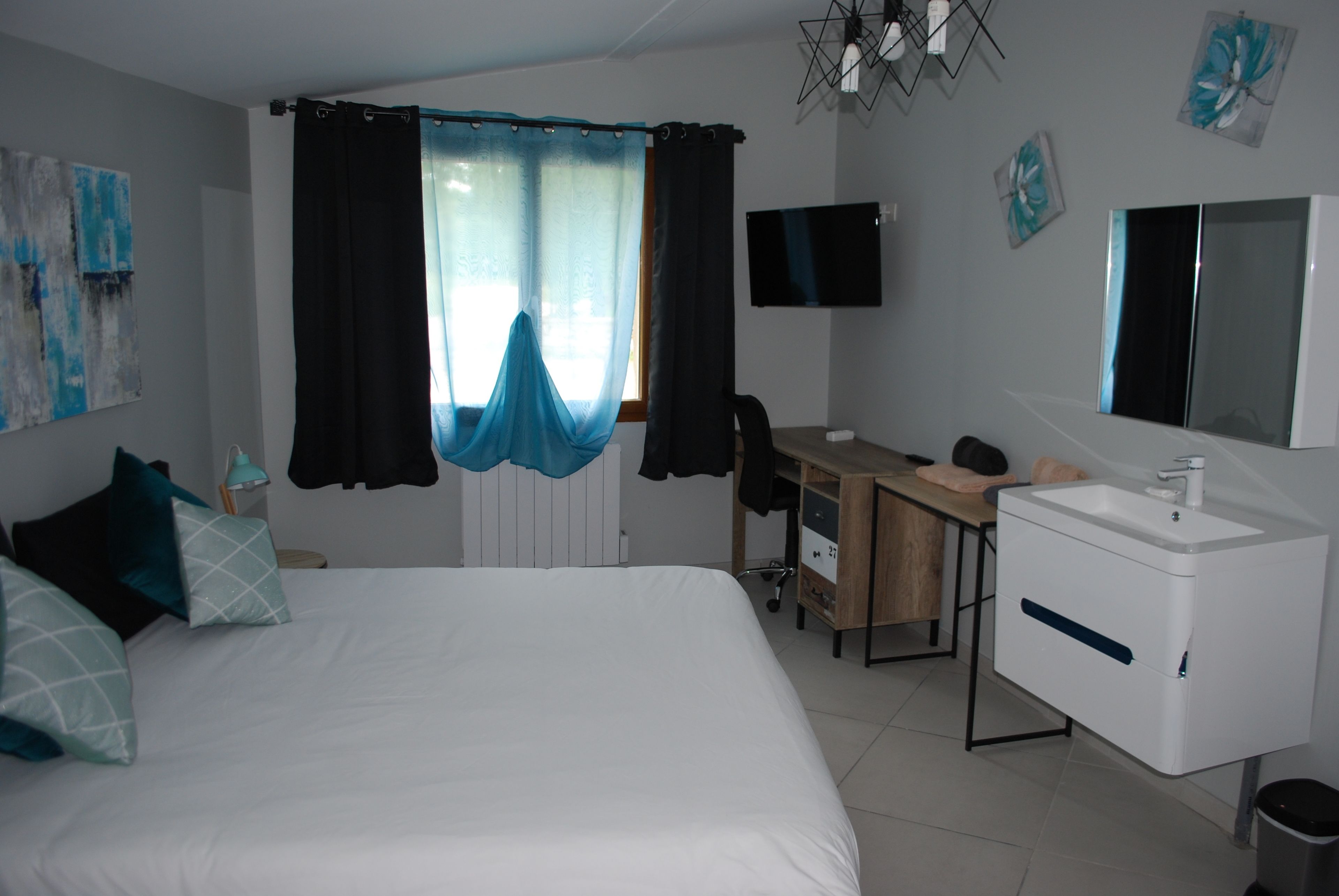 A bedroom with a bed, a desk, a chair, a TV, and a bathroom with a sink and mirror.