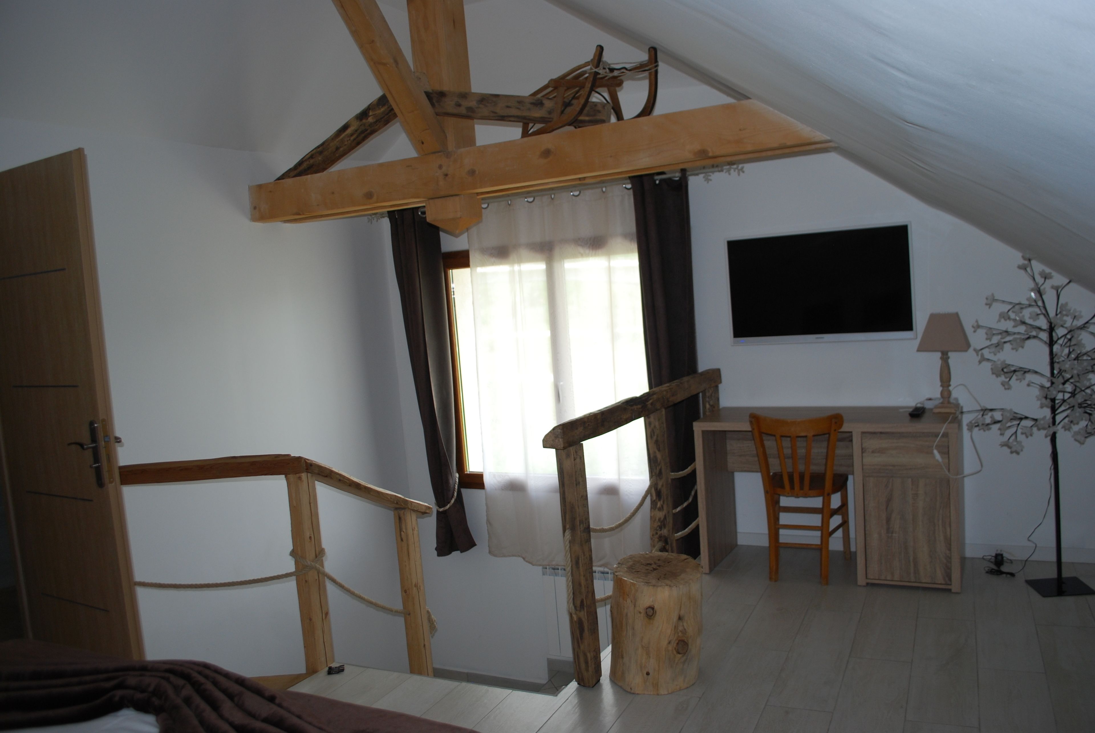 Une pièce avec un plafond en pente, des poutres en bois, un bureau avec une chaise, un petit tabouret et une plante.