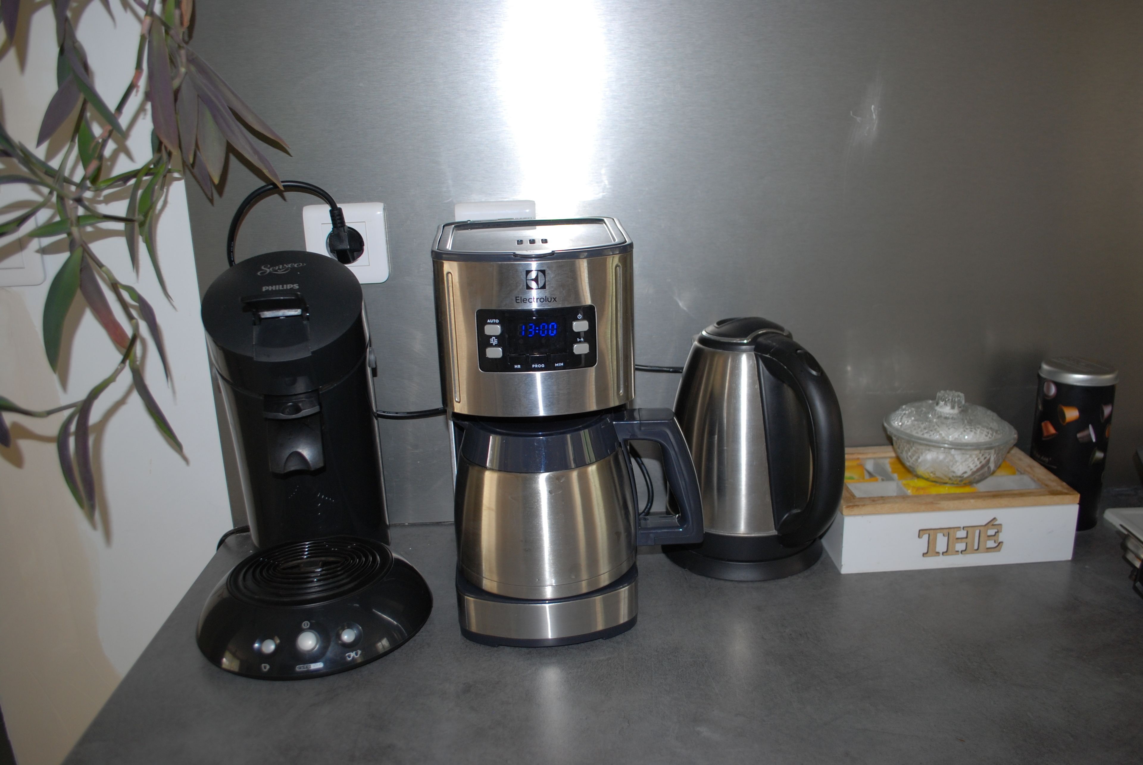 Eine Kaffeemaschine, ein Wasserkocher und eine mit 'THÉ' beschriftete Schachtel auf einer Küchenarbeitsplatte.