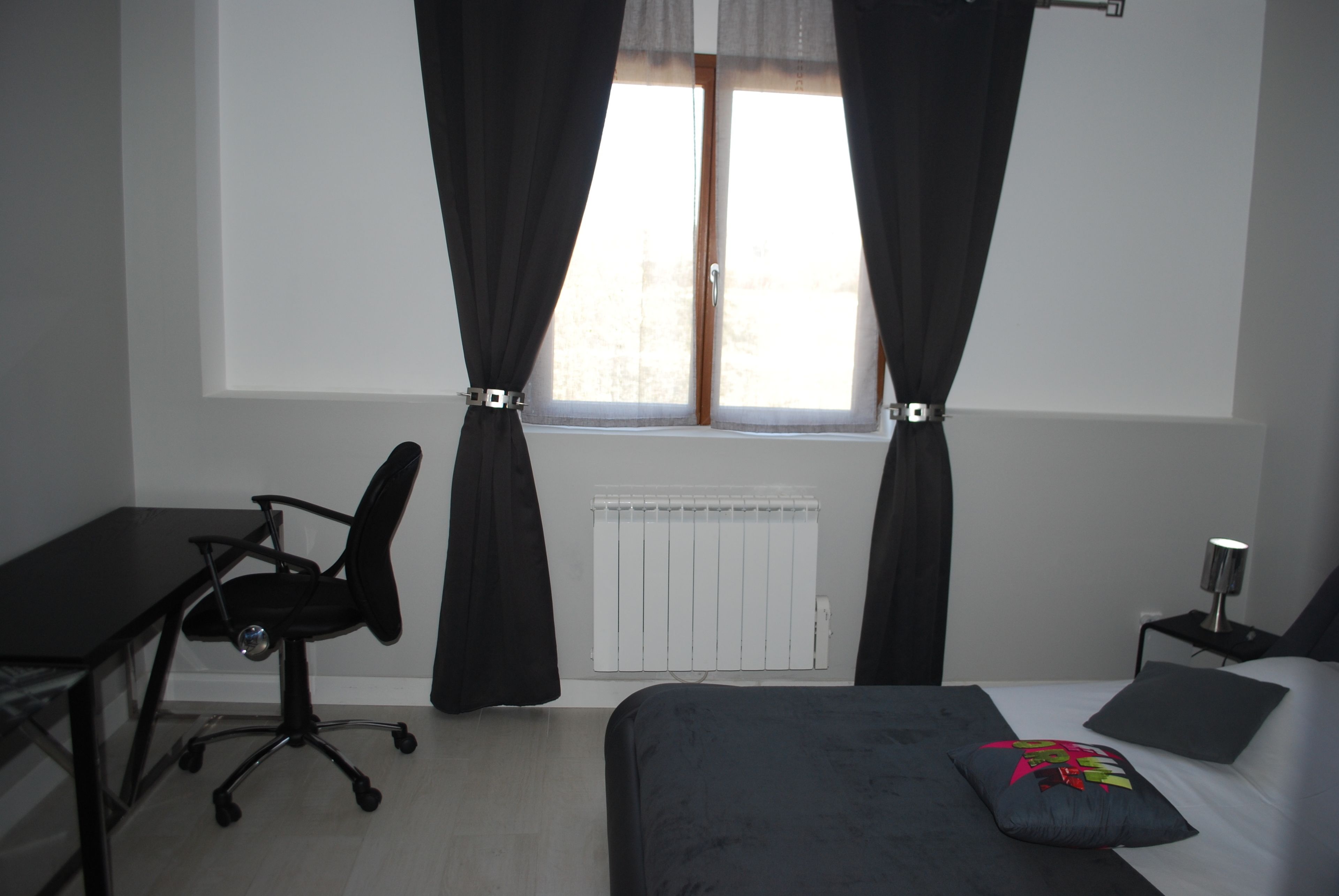 Un dormitorio con cama, escritorio, silla, radiador y ventana con cortinas.