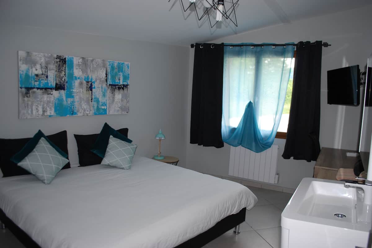 Un dormitorio con cama, mesita de noche, lámpara, ventana con cortinas, televisor instalado en la pared y baño con lavamanos.