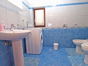 A bathroom with a washing machine, two sinks, a toilet, and a window with a curtain.