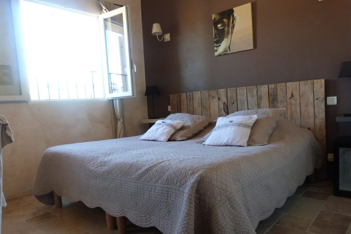 A bedroom with a wooden headboard, a large bed with a quilted bedspread, and a window with blinds.