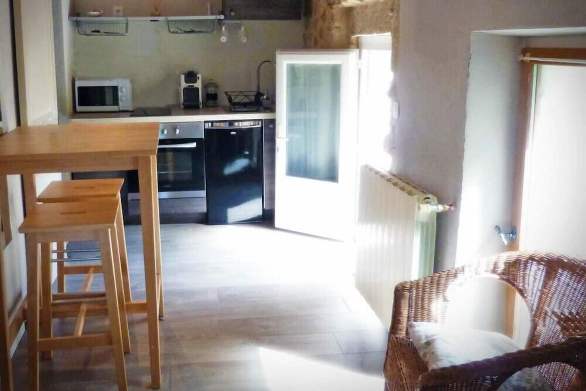 A compact kitchen with a microwave, toaster, and dishwasher. There's a wooden table and a wicker chair.