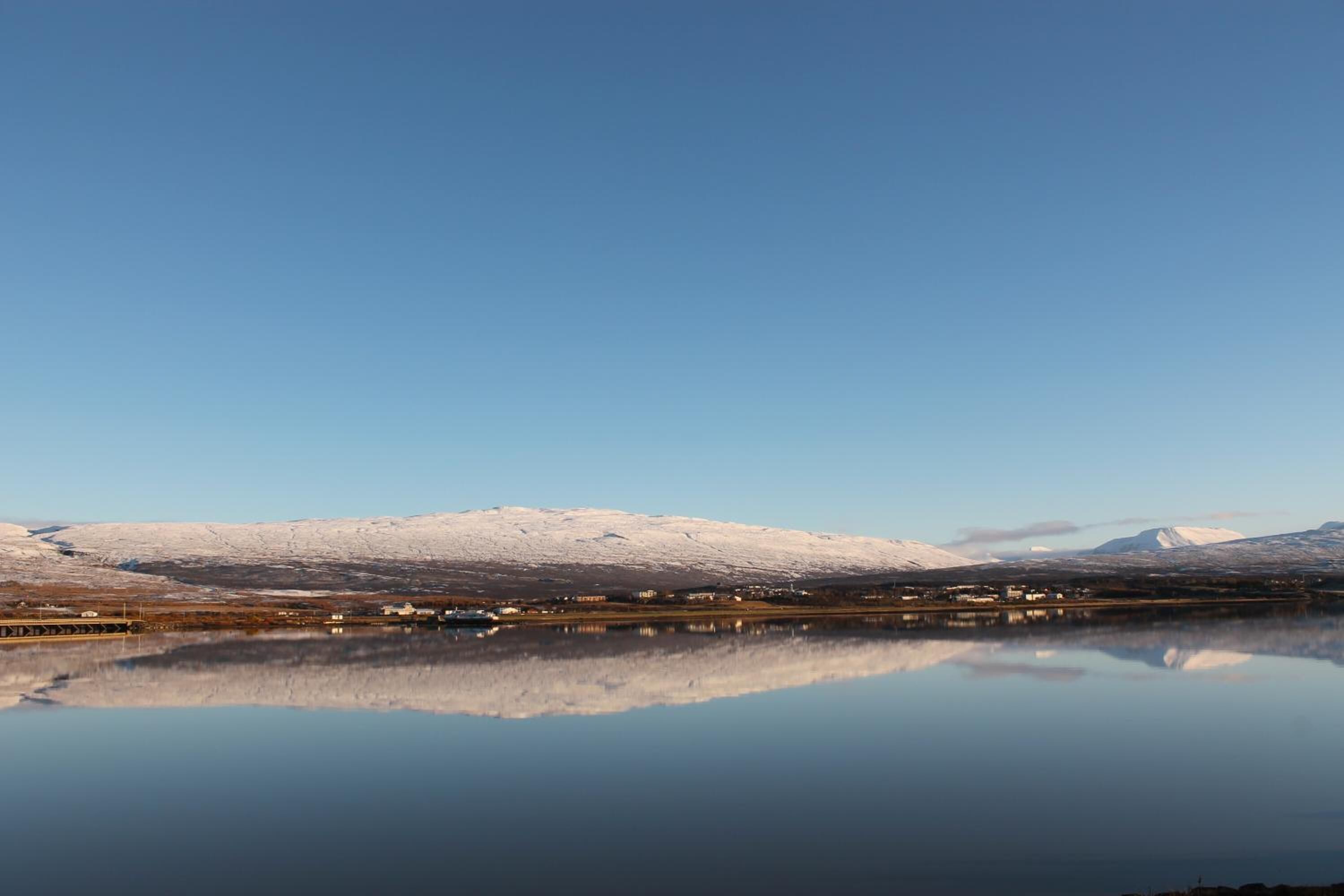 A calm lake reflecting snow-covered mountains and a small settlement with buildings along the shore.