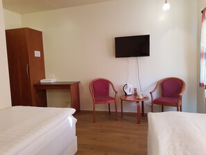 A hotel room with two single beds, a wooden wardrobe, a small desk, a television mounted on the wall, and two red patterned chairs around a small table with a kettle and cups. - Skipalækur Guesthouse (Egilsstaðir)