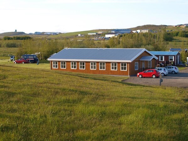 A single-story building with a sloped roof, surrounded by a grassy field and parked cars. - Skipalækur Guesthouse (Egilsstaðir)