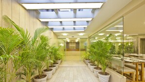 A corridor with potted plants, a glass wall, and a seating area with tables and chairs. - Scenaria Hotel (Ahmedabad)