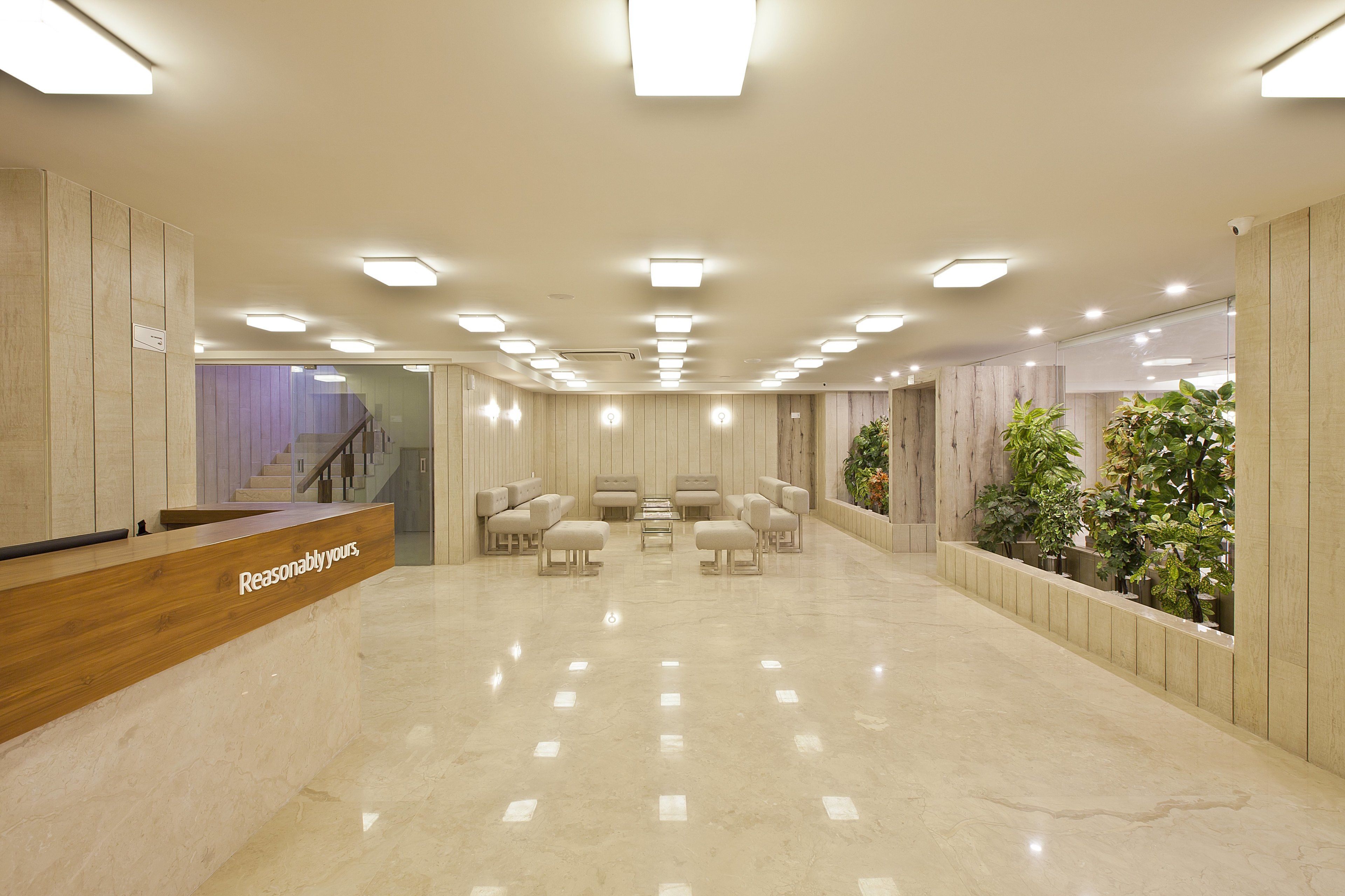 A reception area with a wooden desk labeled "Reasonably Yours," a seating area with chairs and ottomans, and a plant wall on the right.