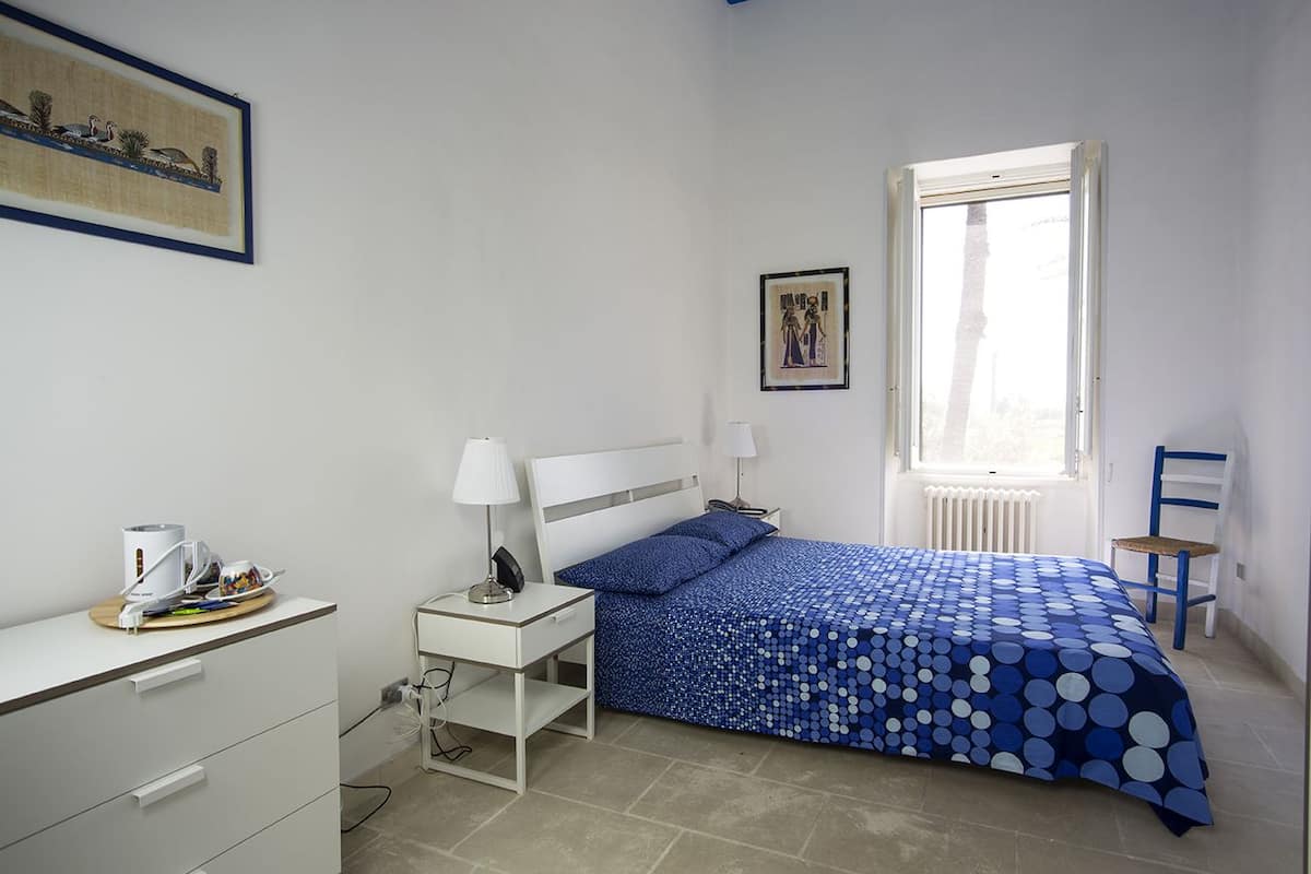 Ein Schlafzimmer mit Bett, Nachttischen, Schreibtisch, Stuhl, Fenster und Bildern an den Wänden.
