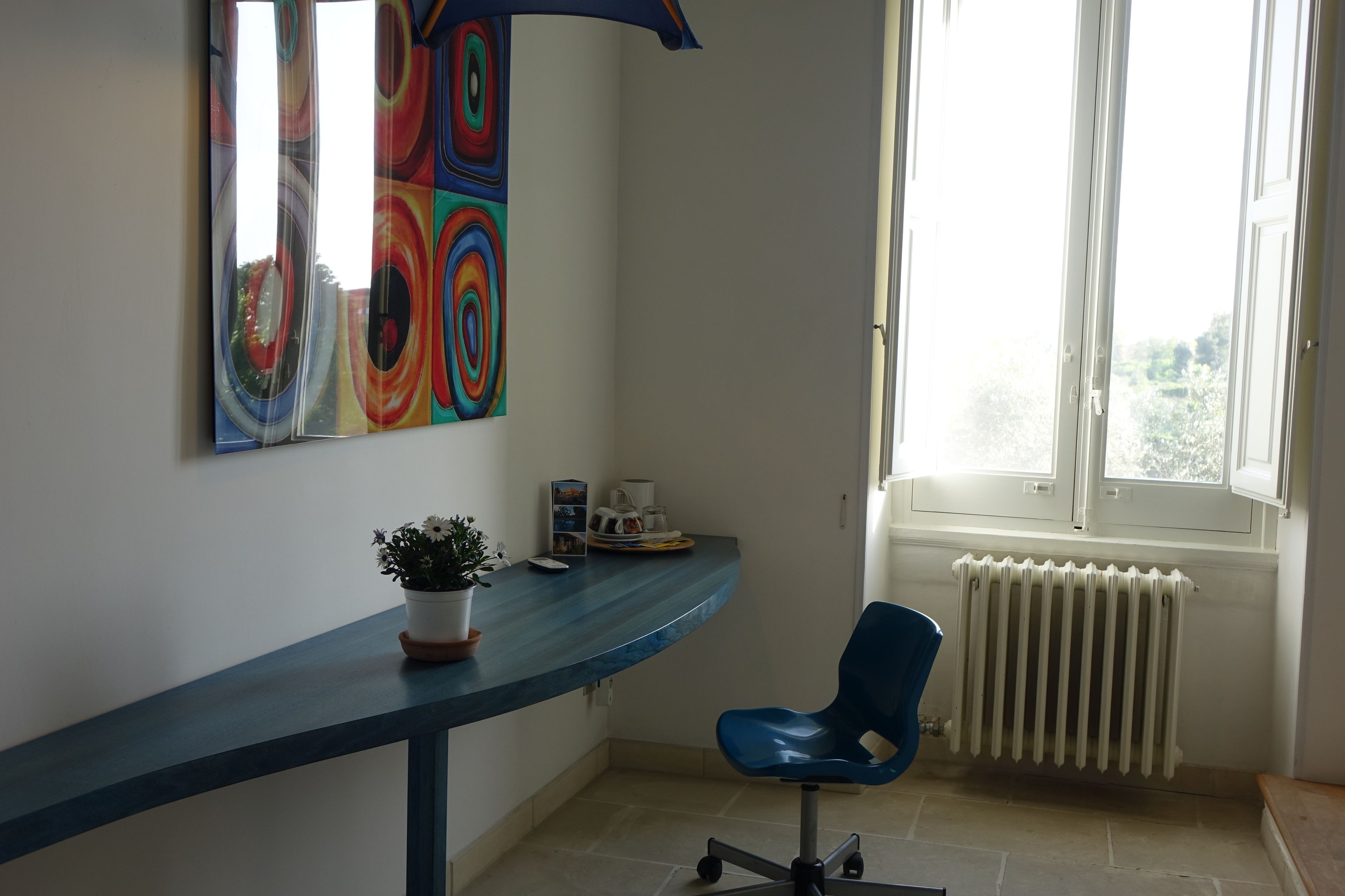 A room with a curved blue desk, a blue office chair, a radiator, and a window with a view of greenery.