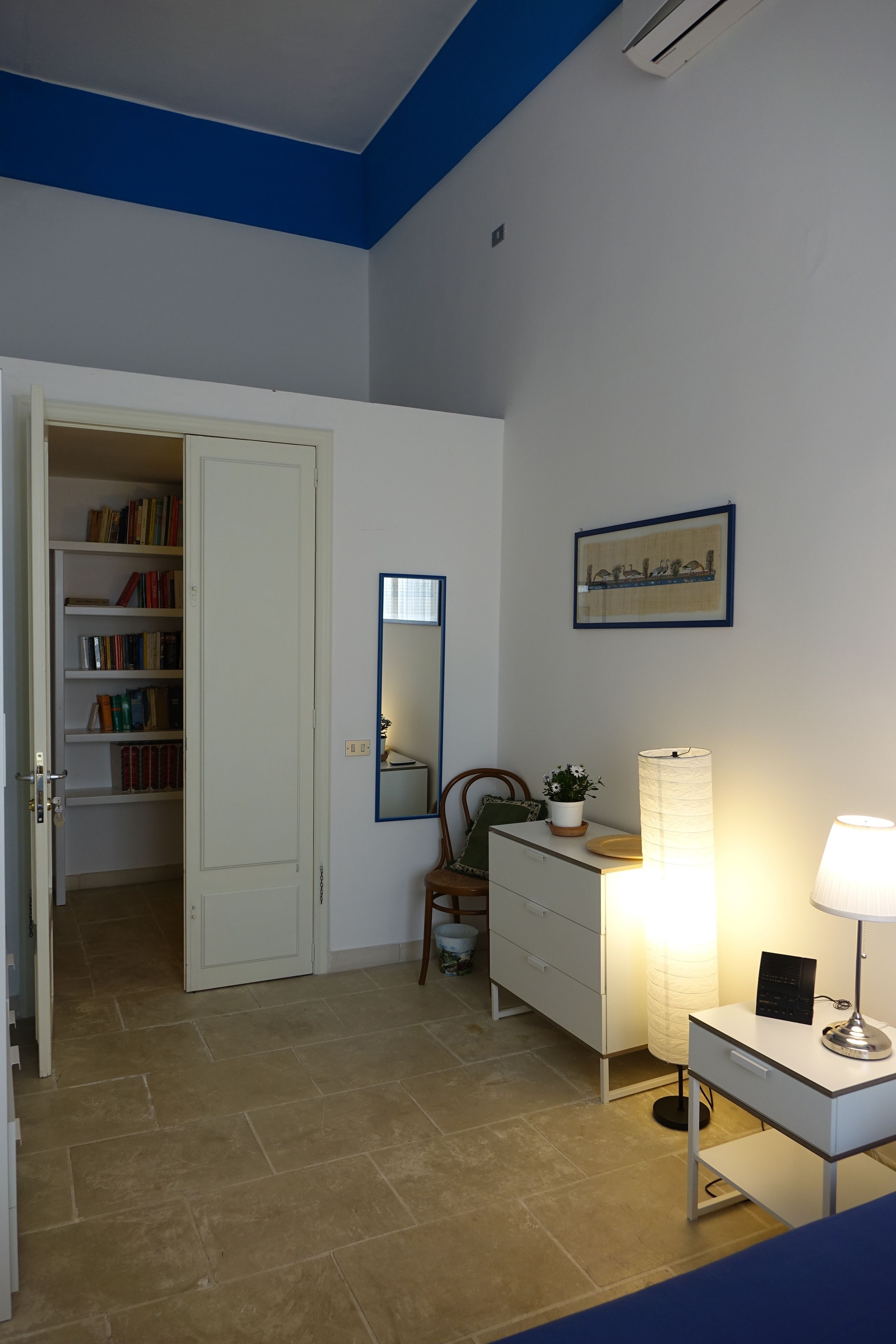 Ein Zimmer mit einem weißen Kleiderschrank, einem Schreibtisch mit Lampe, einem Stuhl und einem Bücherregal voller Bücher.