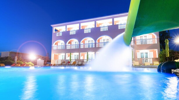 Ein Schwimmbecken mit einer Springbrunnenanlage, ein mehrstöckiges Gebäude mit Balkonen im Hintergrund und ein klarer Himmel.
