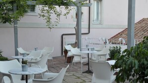 An outdoor seating area with white plastic chairs and tables, potted plants, and a pink building in the background. - Hotel Villa Pimpina (Carloforte)