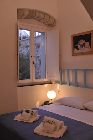 Ein Schlafzimmer mit Bett, Nachttisch, Lampe und einem Fenster mit Blick auf Grünflächen.