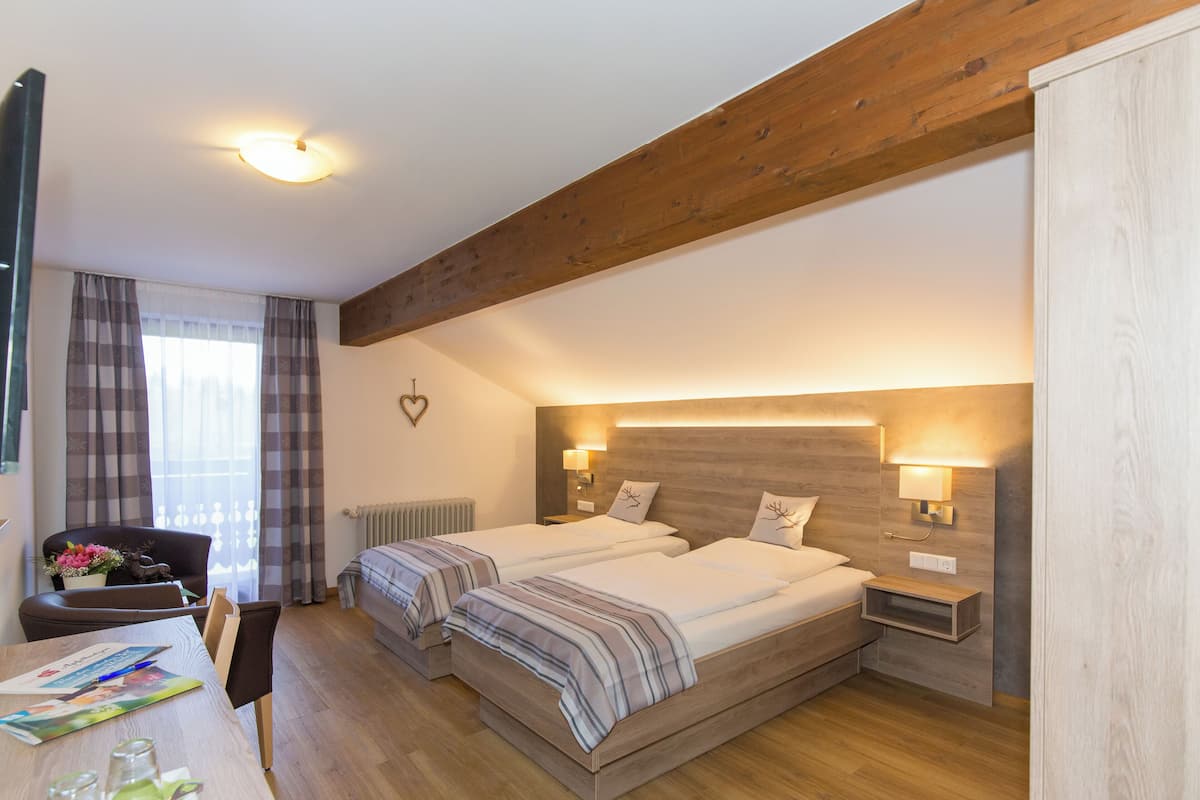 A bedroom with two beds, a wooden ceiling, a nightstand with a lamp, a desk with a chair, and a window with curtains.