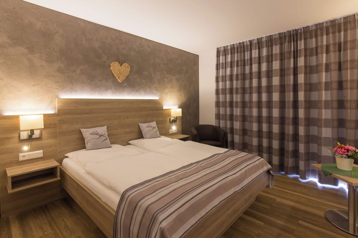 A hotel room with a large bed, bedside lamps, a heart-shaped wall decoration, and a view of the outdoors through curtains.