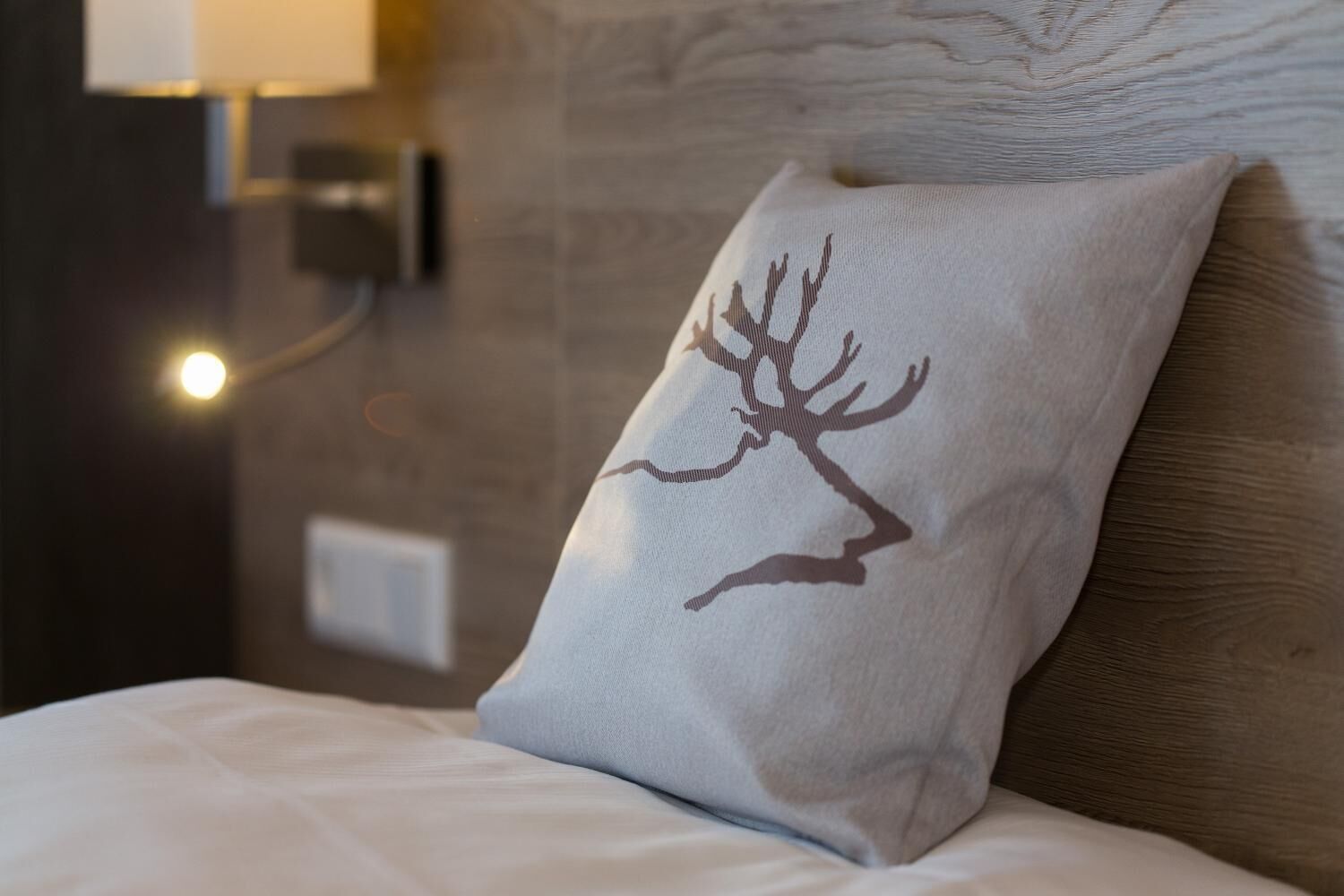 A bed with a pillow featuring a deer silhouette, a wall-mounted light, and a switch.