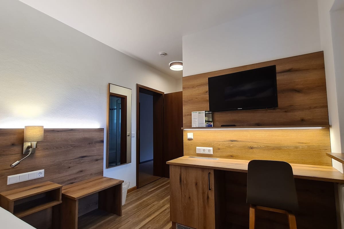 A modern hotel room with a wooden accent wall, a flat-screen TV, a desk with a chair, and a small seating area.
