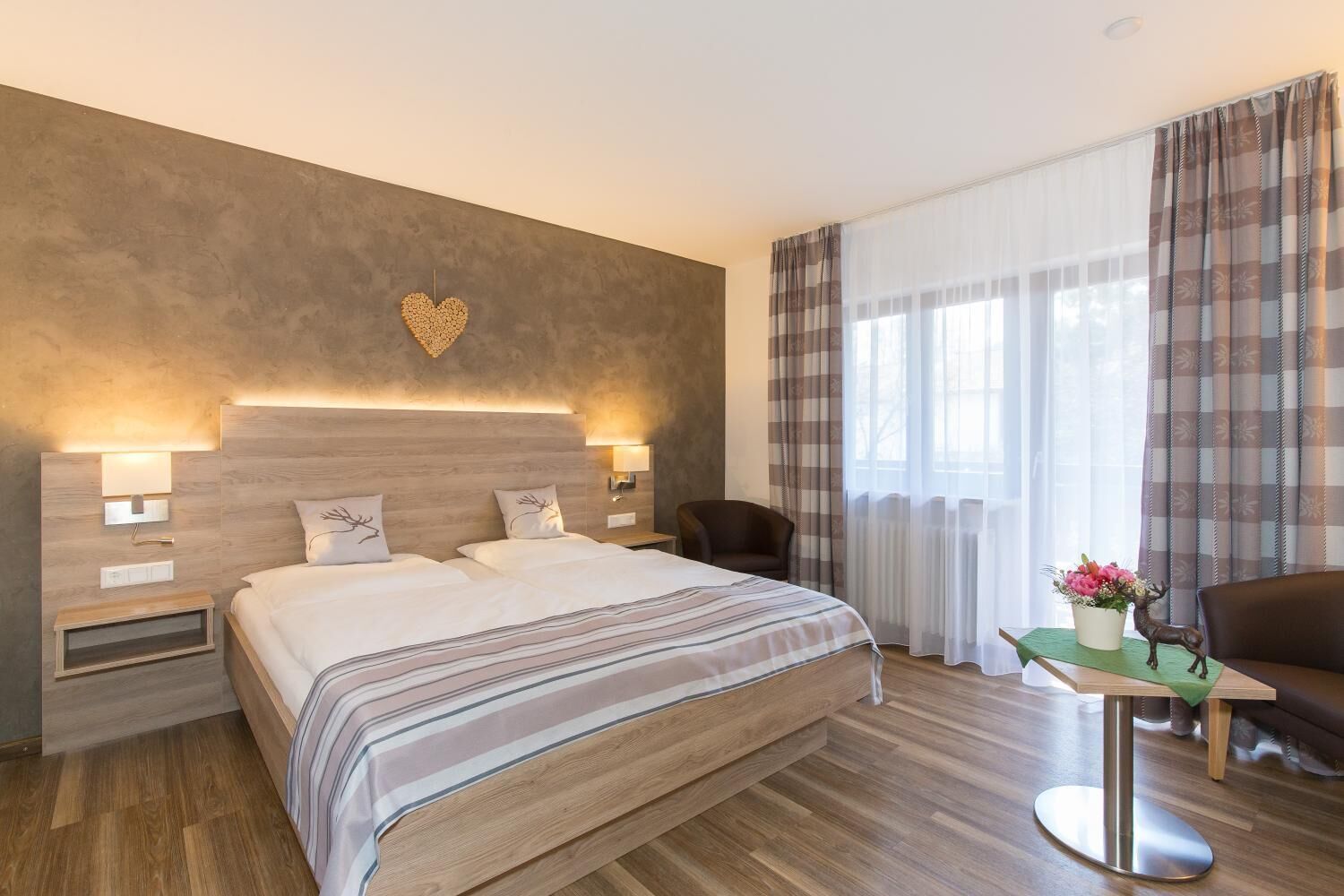 Ein Hotelzimmer mit einem großen Bett, Nachttischlampen, einer Wanddekoration in Herzform, einem Fenster mit Vorhängen und einem kleinen Tisch mit Blumenvase und einer Hirschfigur.