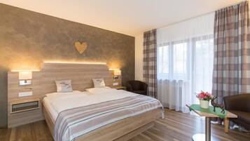 Ein Hotelzimmer mit einem großen Bett, Nachttischlampen, einer Wanddekoration in Herzform, einem Fenster mit Vorhängen und einem kleinen Tisch mit Blumenvase und einer Hirschfigur.