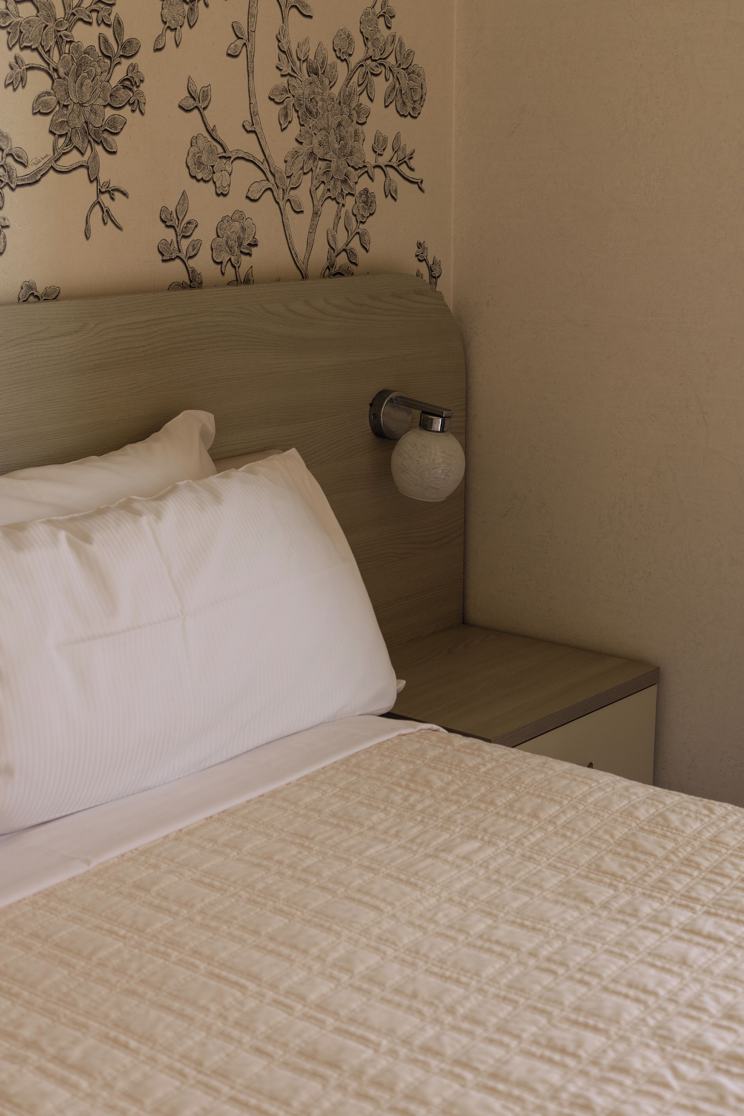 Een net opgemaakt bed met een gewatteerde spreideken, een nachtkastje met een lamp, en een wand met bloemenbehang.