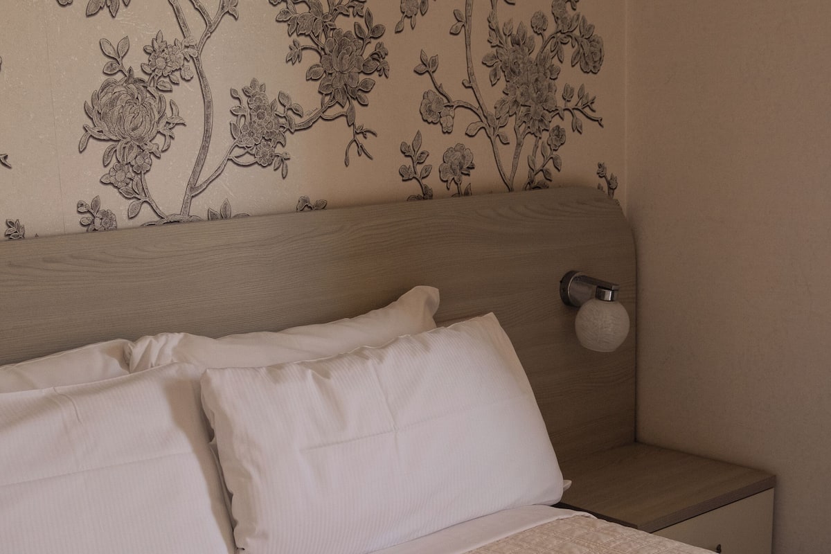 Una camera da letto con un letto, due cuscini, un comodino e una parete con carta da parati a motivi floreali.